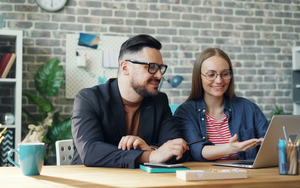 Male And Female Coworkers Are Discussing Work Sitting At Desk With Laptop Together Talking Smiling. Cooperation, Business People And Communication Concep