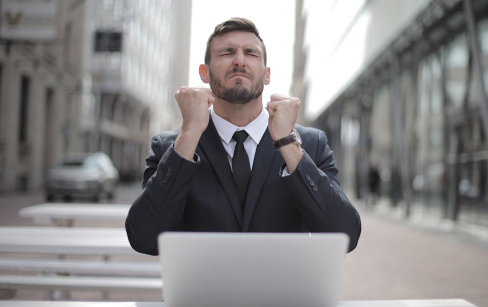 Businessman Sitting Outdoors in Front of a Laptop, Celebrating Success with Clenched Fists and a Triumphant Expression